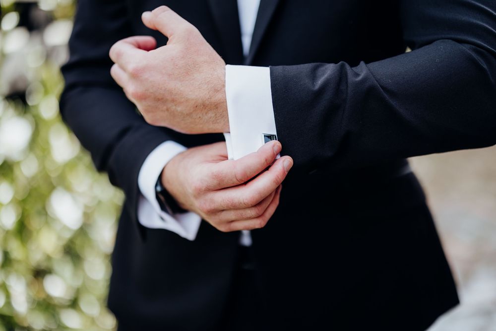 Groom in black tuxedo adjusting silver cufflinks on white dress shirt sleeves outdoors.