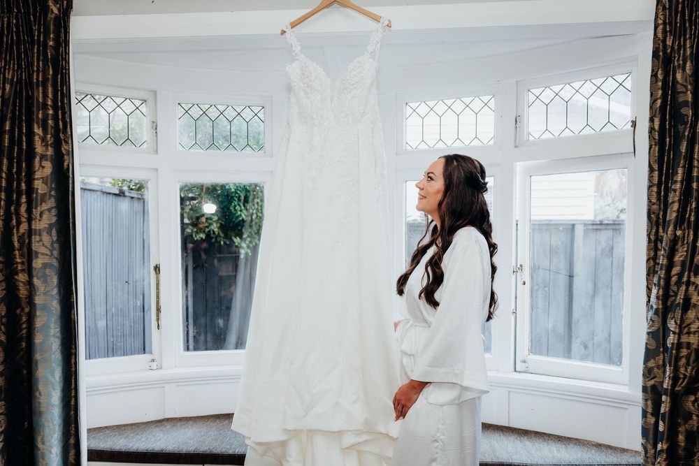 Bride in white robe admiring her elegant wedding dress hanging by a sunlit window.
