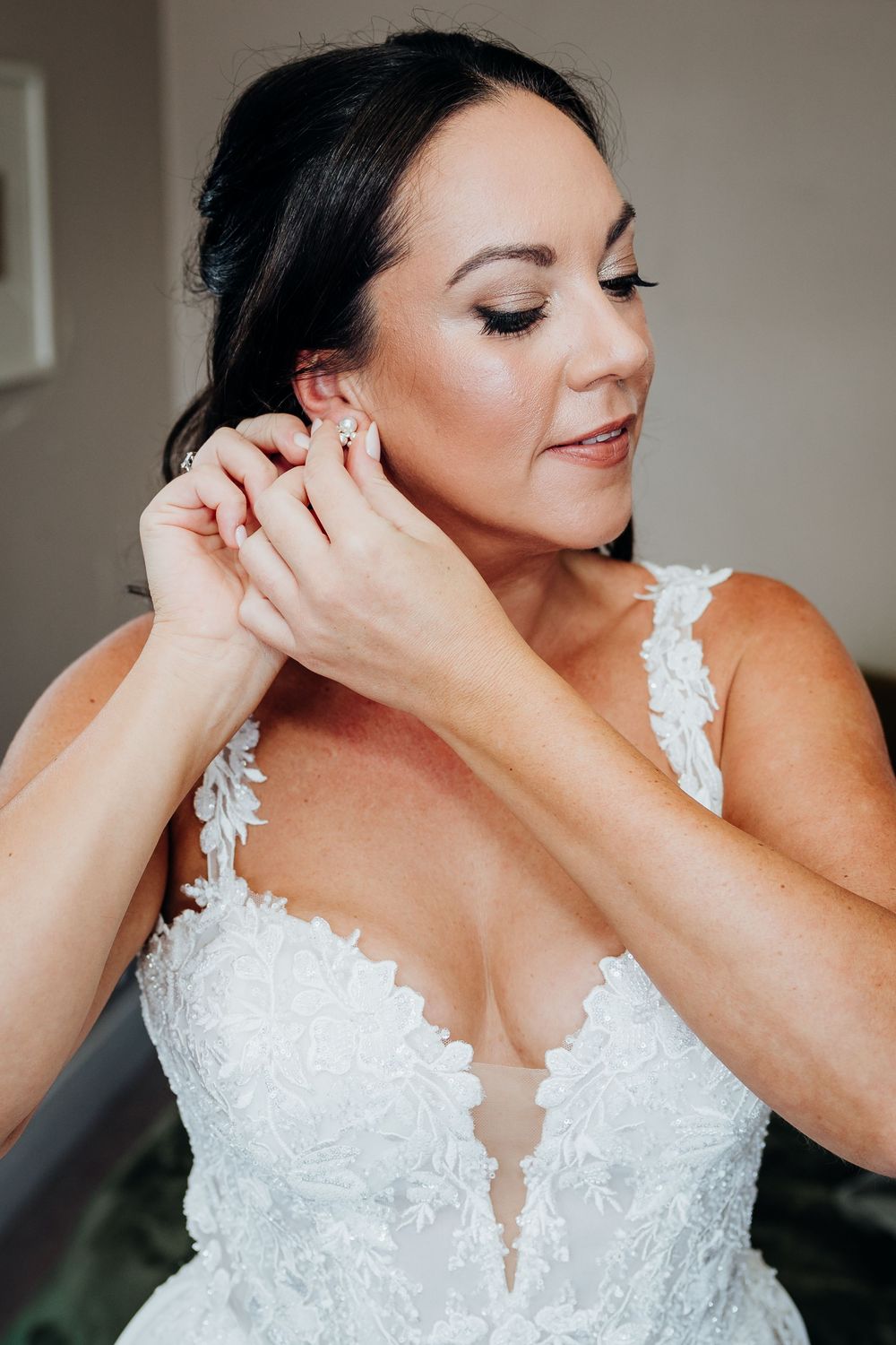 Bride in lace wedding gown putting on earrings while getting ready for her wedding day.