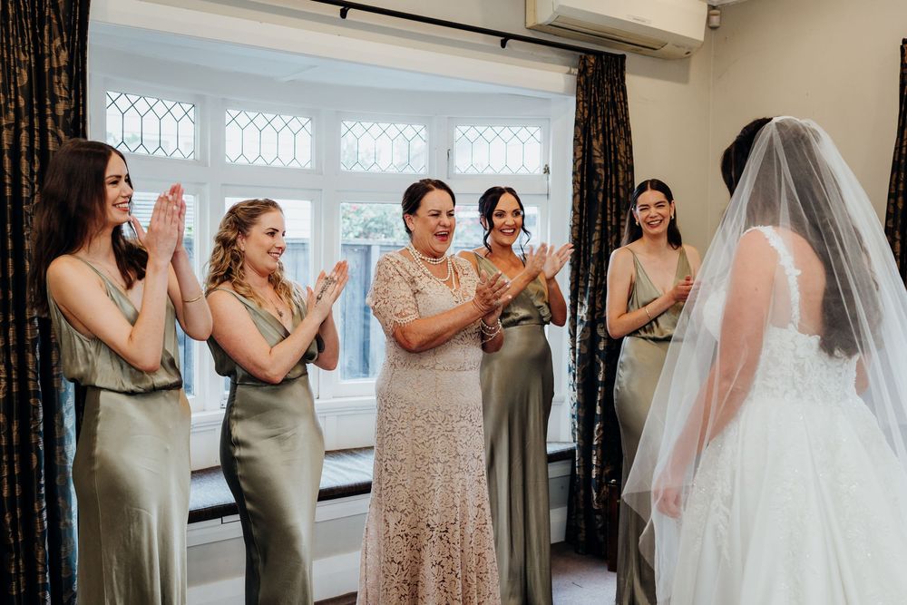 Bridesmaids in gold dresses clapping for bride in white gown and veil during wedding preparations indoors.
