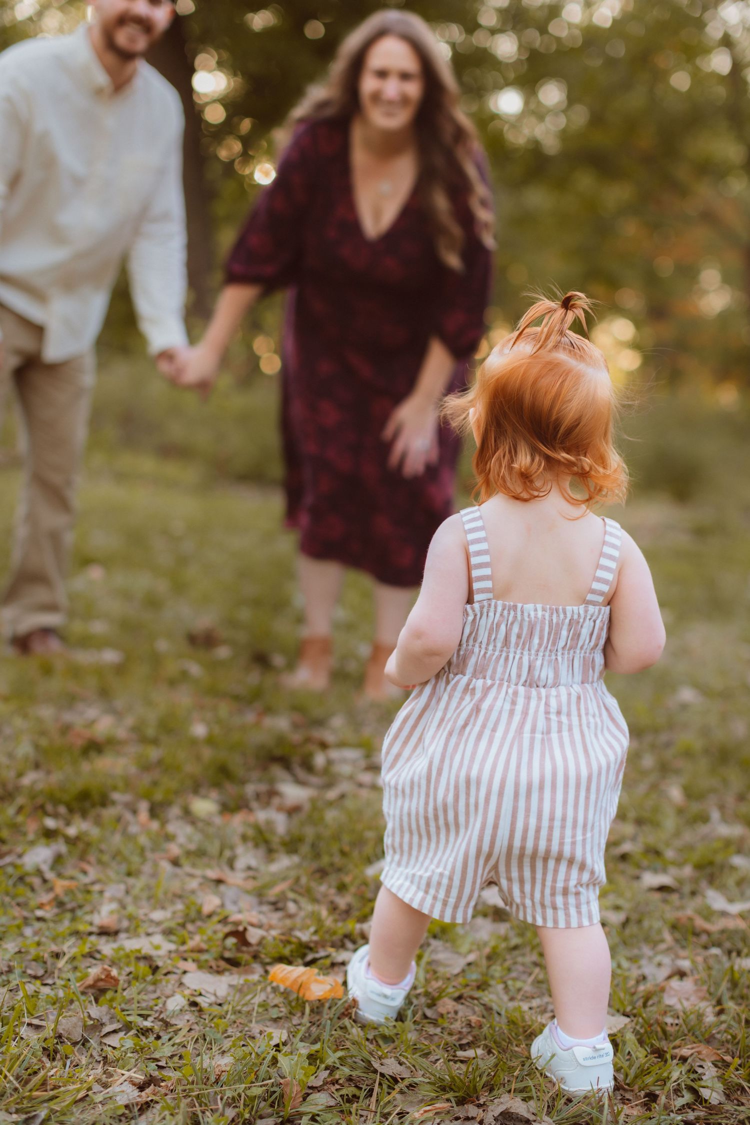 A toddler girl with red hair and striped overalls runs toward her parents' open arms in a field at golden hour.