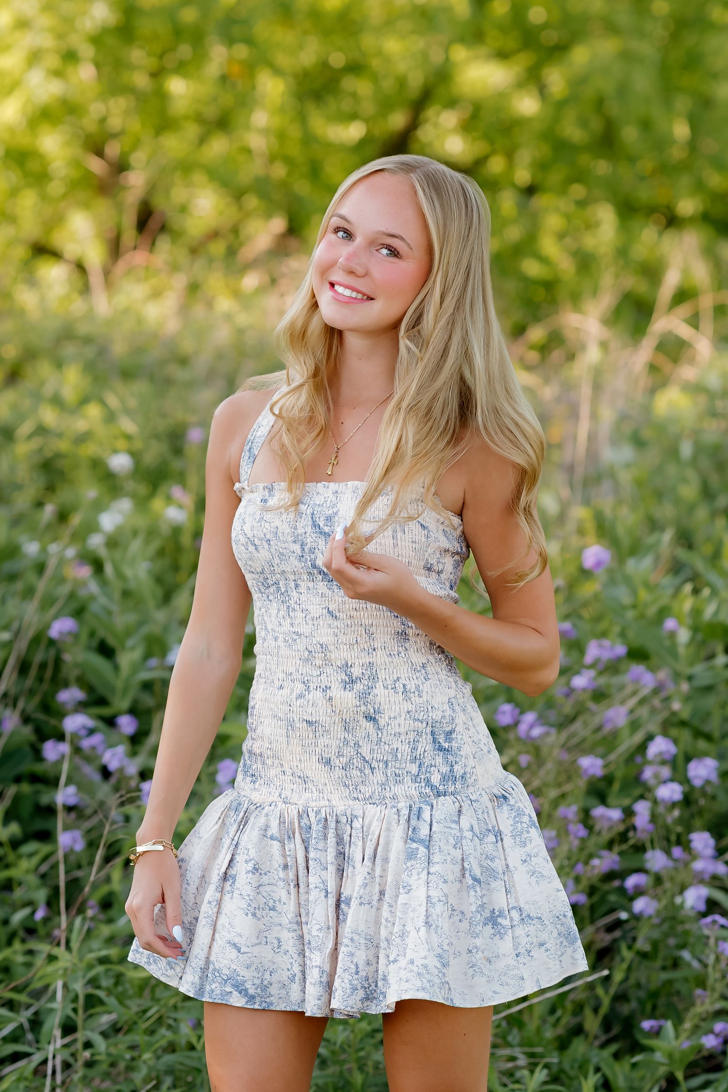 Young blonde woman in floral sundress smiling outdoors among wildflowers and lush green foliage.