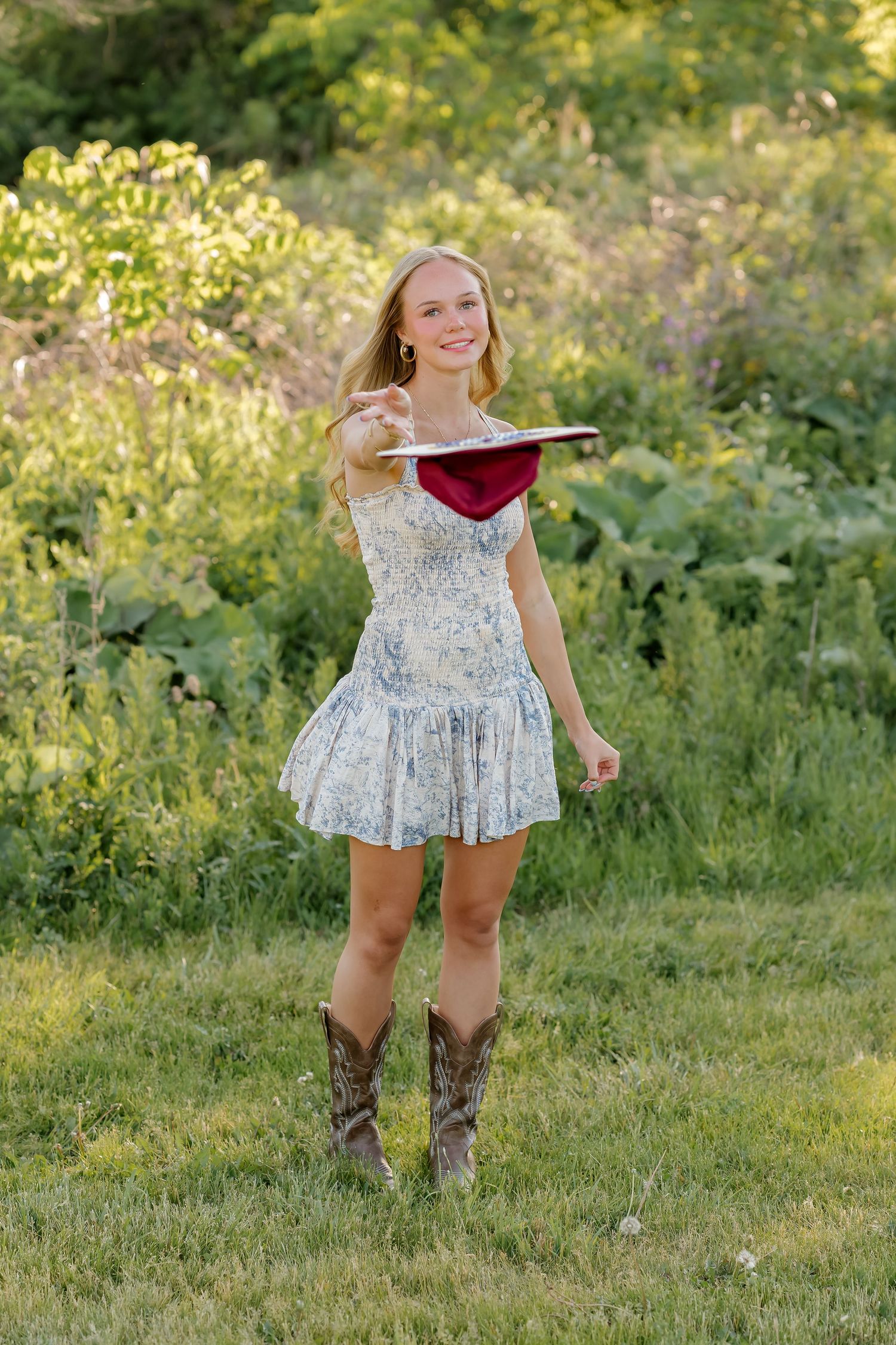 Smiling teenage girl in floral dress and cowboy boots holding a graduation cap outdoors in a green field.