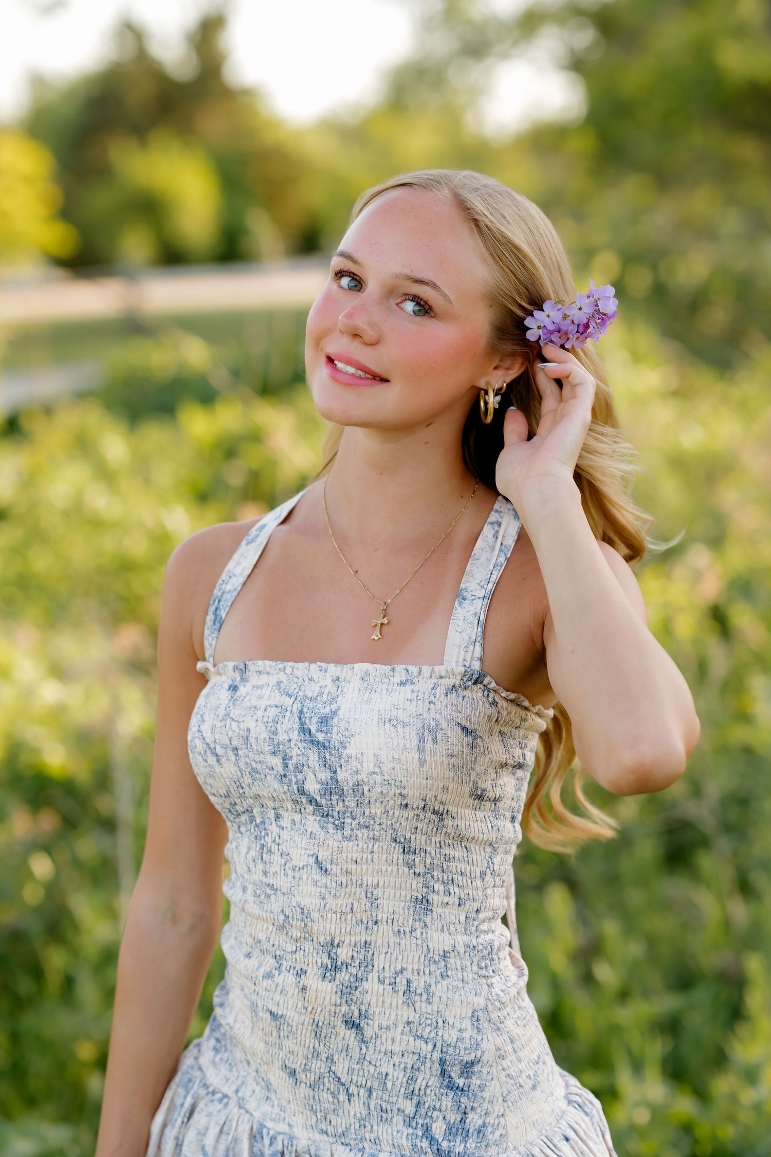 Young blonde woman in blue floral dress holding purple flowers in her hair outdoors.
