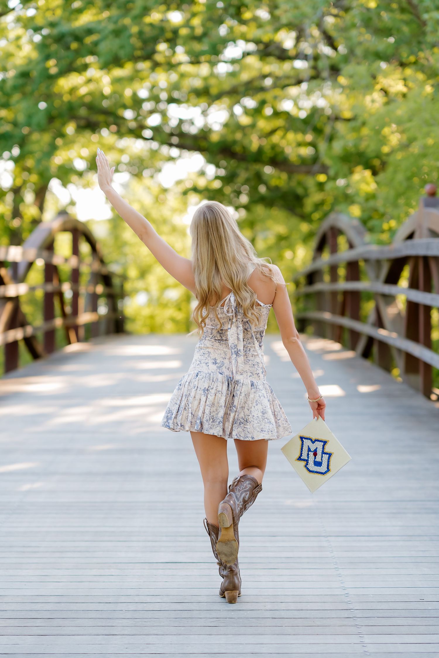 Blonde woman in floral mini dress and cowboy boots walks on wooden bridge holding MU hat with arm raised.