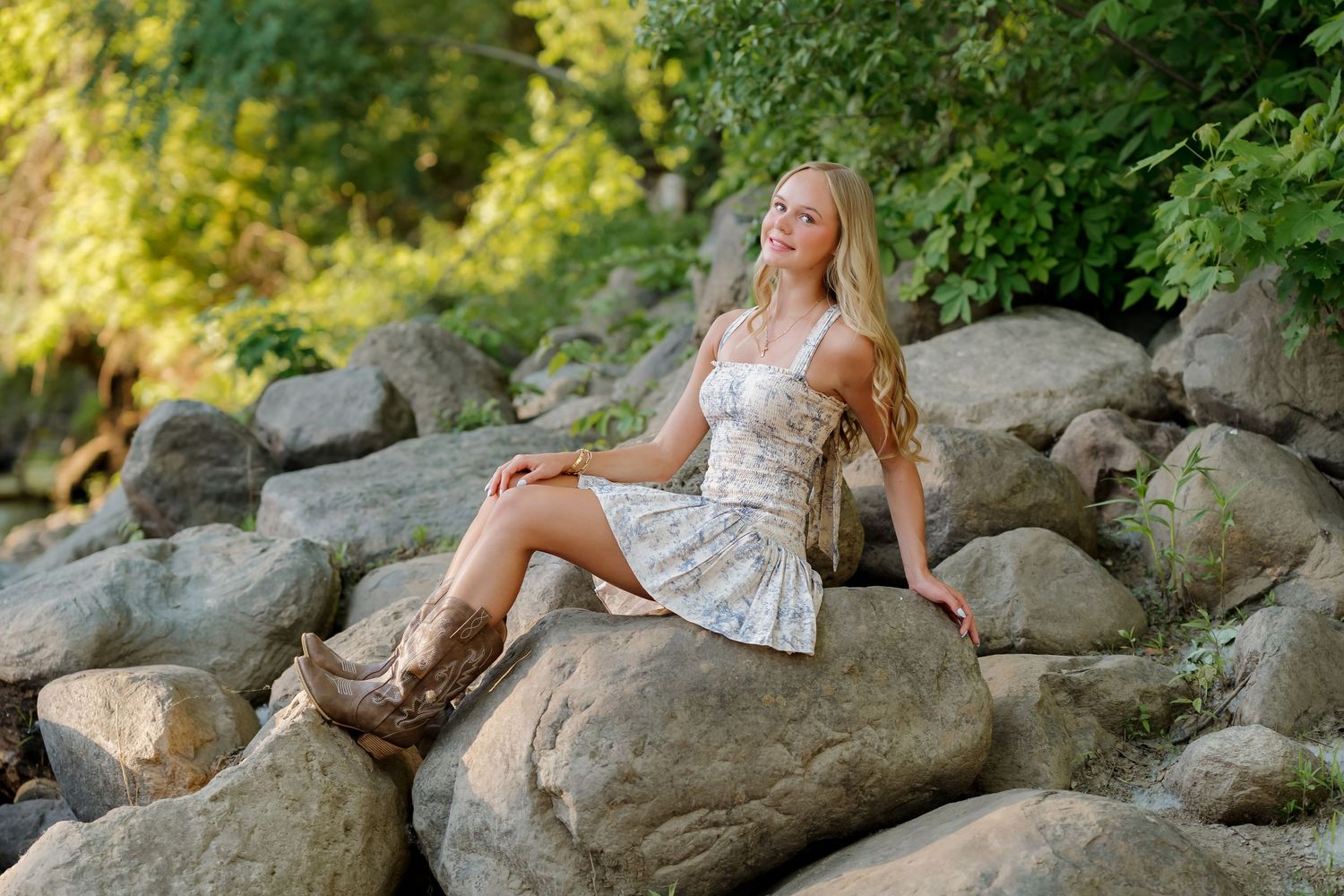 Young woman in floral dress and cowboy boots sitting on rocks surrounded by lush green trees.