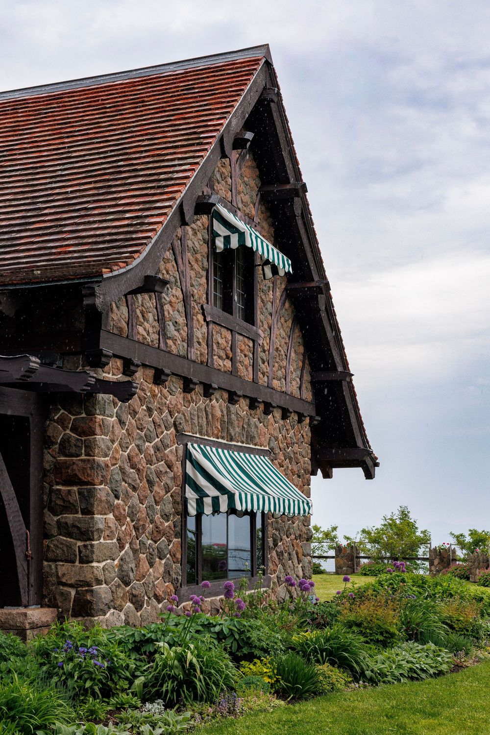 Stone Tudor-style building with green striped awnings, red tile roof, and lush garden landscaping under cloudy sky.