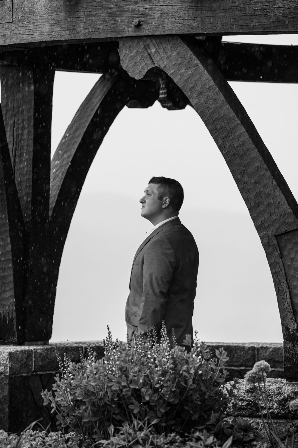 Groom in suit stands under wooden arch beams in black and white wedding portrait with garden flowers.