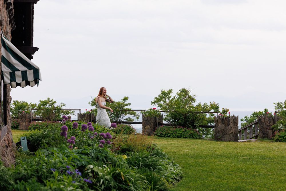 Woman in white dress standing by a wooden fence in a lush garden with purple flowers and ocean views.