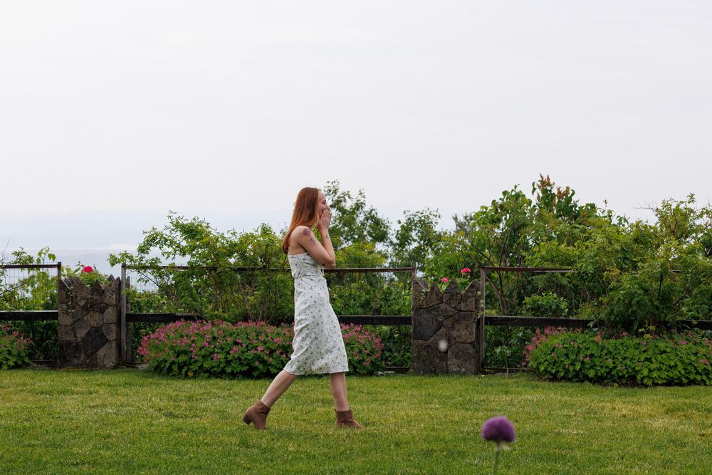 Woman in floral dress walking through lush garden with wooden fence and colorful flowers on overcast day.