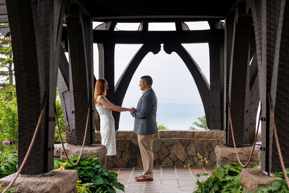 Couple holding hands under a rustic wooden gazebo with scenic ocean views in the background.