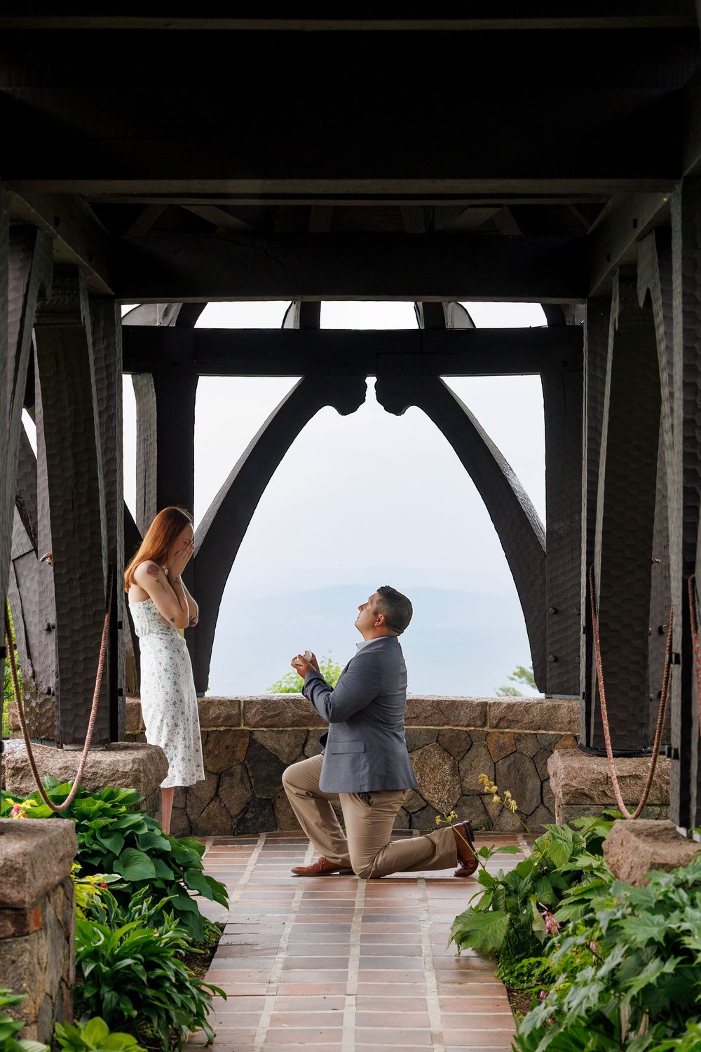 Man proposes on one knee to woman in floral dress under rustic wooden pavilion with scenic water views.