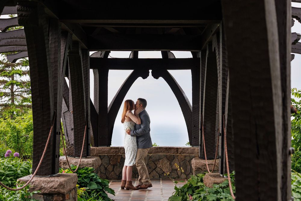 Romantic couple embracing under a dark wooden pavilion with lush greenery and ocean backdrop.