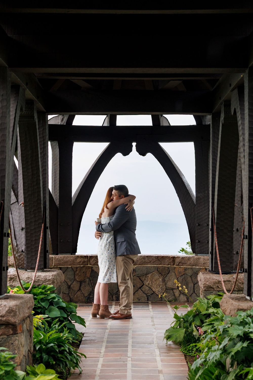 Couple embraces and kisses under a rustic dark wooden pavilion with scenic ocean views in background.