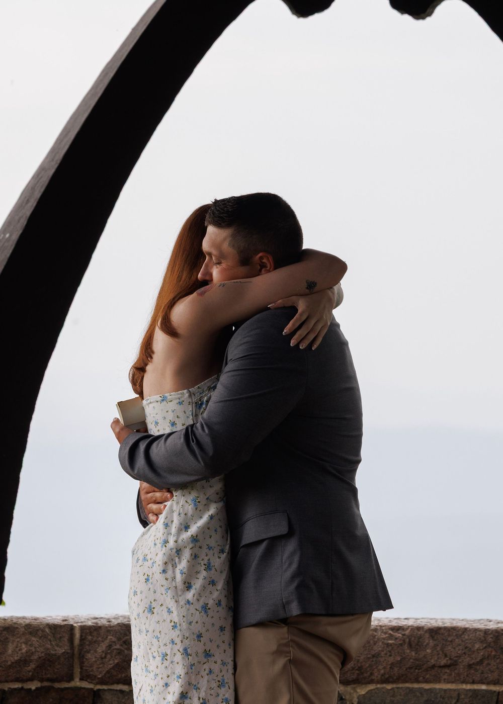 Couple sharing a romantic embrace under a stone arch with an ocean view in the background.