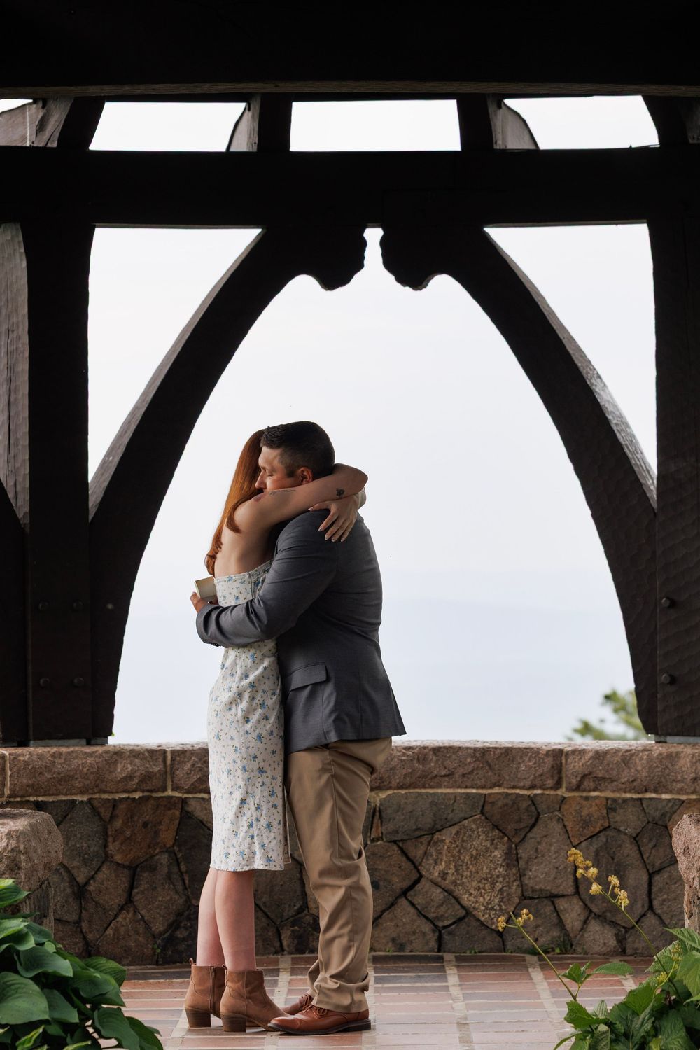 Couple embracing under rustic wooden arch pavilion with stone wall and ocean view backdrop.