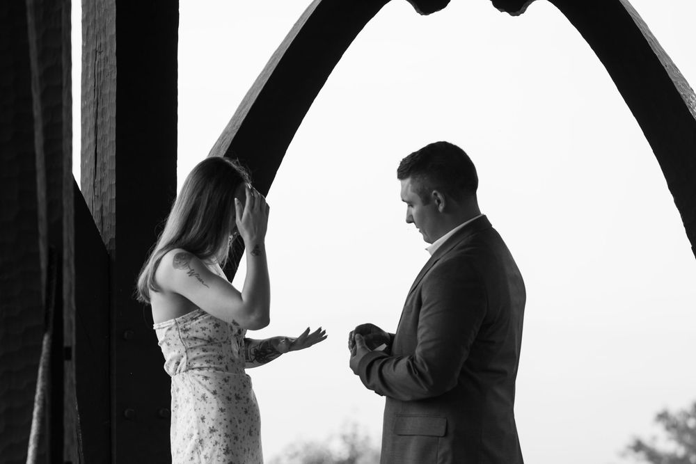 Black and white photo of a man proposing to an emotional woman under a wooden arch structure outdoors.