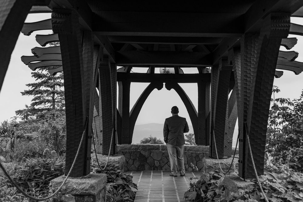 Black and white photo of a man standing alone under a wooden gazebo structure overlooking a scenic view.