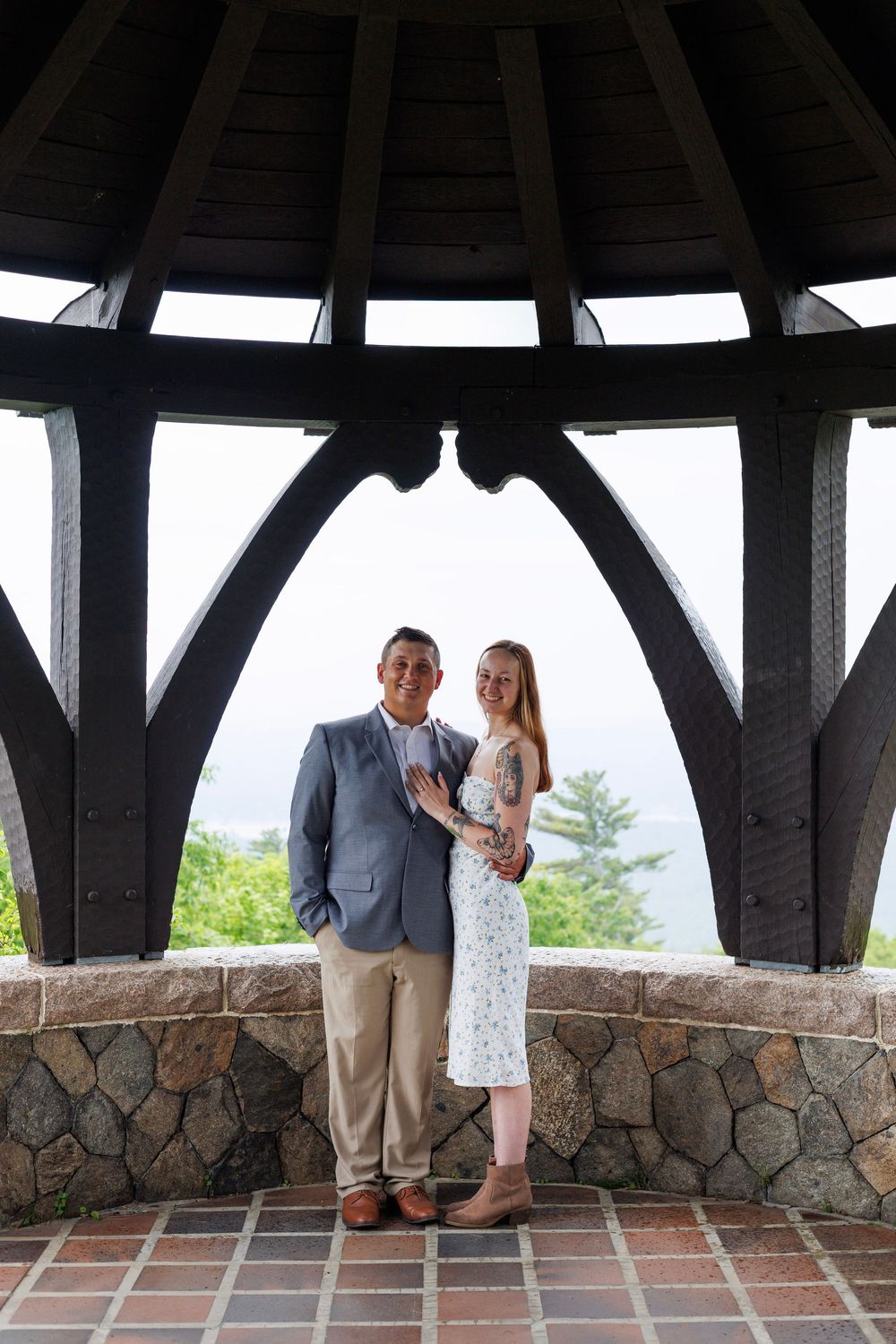 Smiling couple posing together inside a wooden gazebo with a scenic landscape visible behind them.