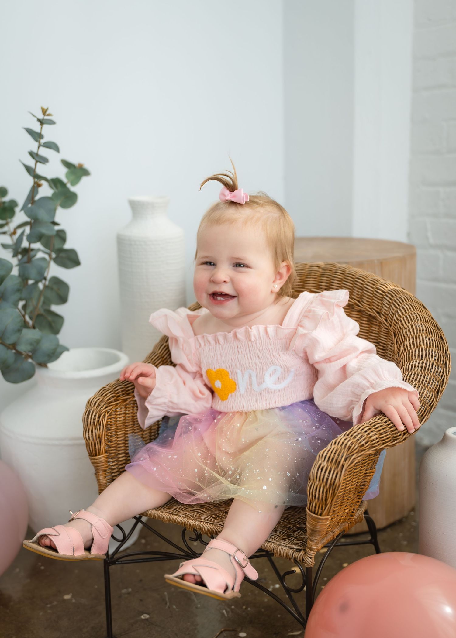 Baby girl in pink ruffle romper with tutu sits smiling in wicker chair, first birthday photo with balloons.