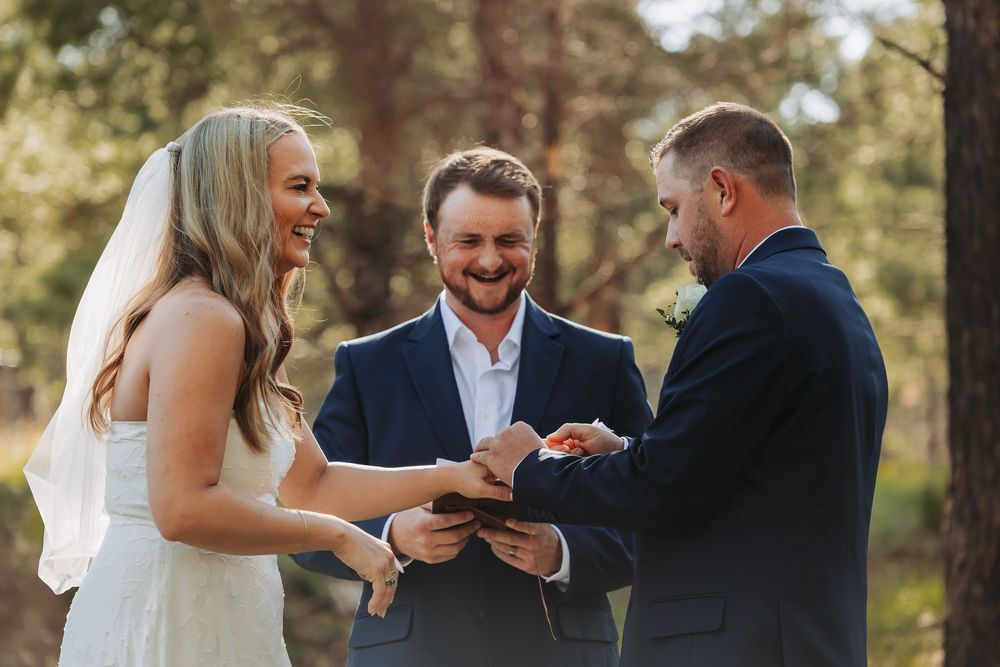 Bride and groom exchange rings during an outdoor forest wedding ceremony, smiling joyfully with their officiant by their side.