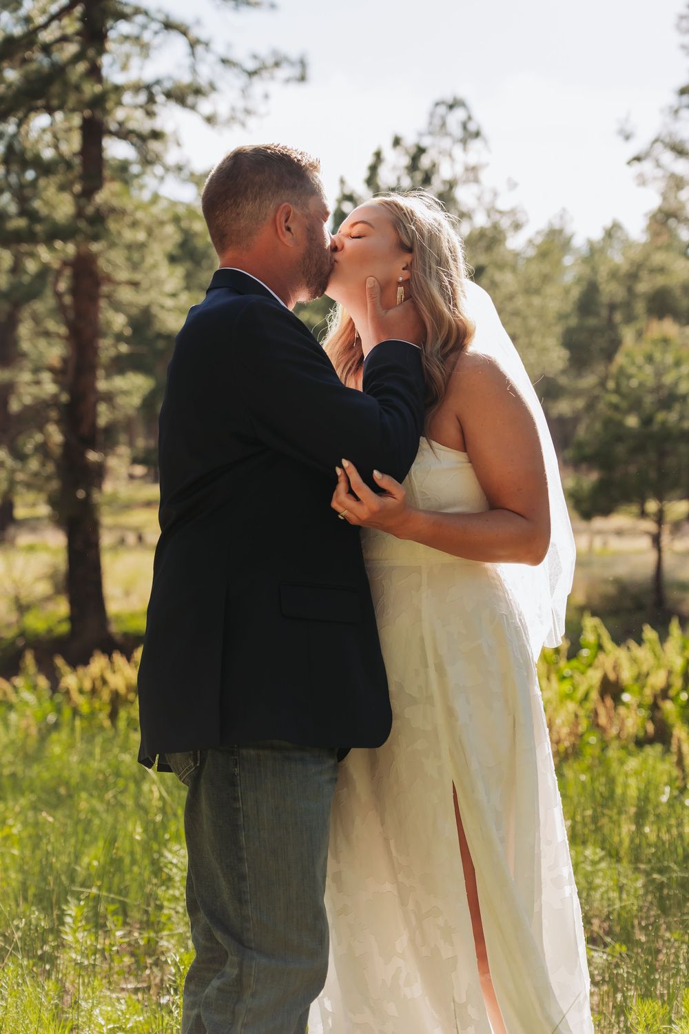 Bride and groom share a romantic kiss outdoors surrounded by lush green trees and meadow on their wedding day.