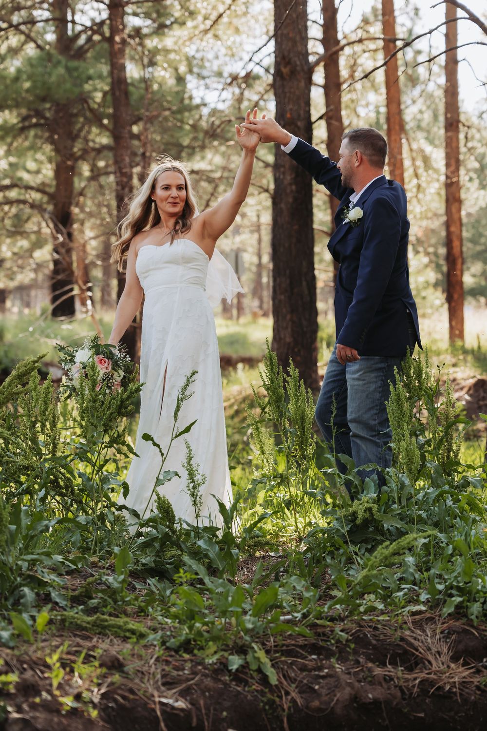 Bride and groom dancing together in a forest setting, bride in white gown holding bouquet as groom spins her.