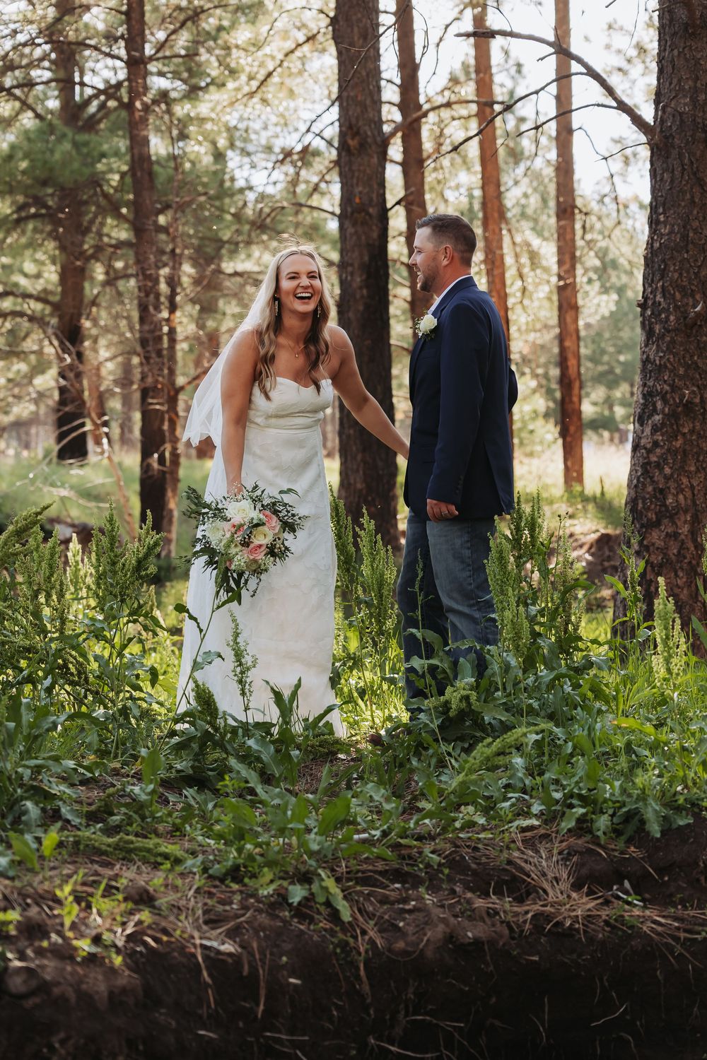 Bride in white gown laughing with groom in navy blazer holding bouquet in sunlit forest wedding photo.