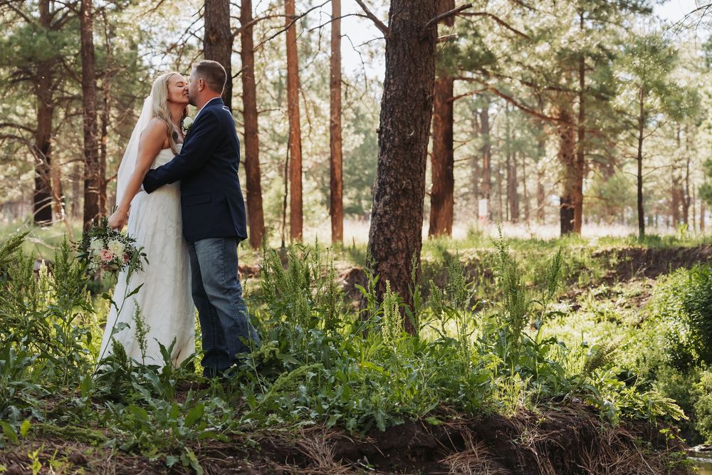 Bride and groom embracing in a lush forest setting, surrounded by tall pine trees and green ferns on their wedding day.