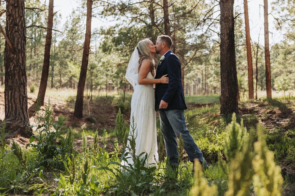 Bride and groom sharing a romantic kiss in a lush forest setting, surrounded by tall pine trees and green foliage.