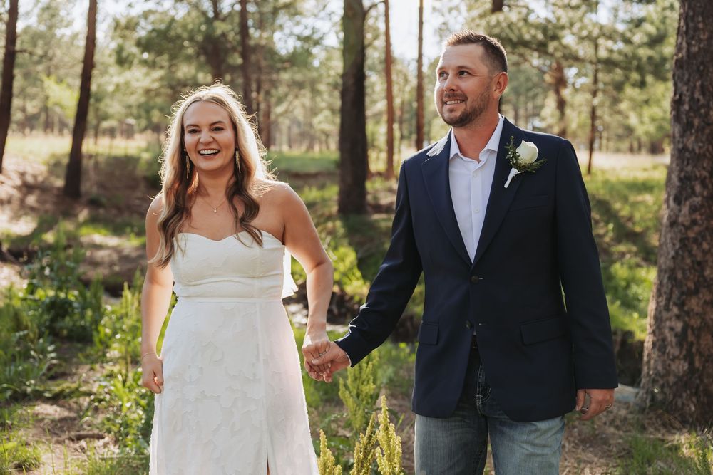 A smiling bride in a white dress and groom in a navy suit hold hands while walking through a sunlit pine forest.