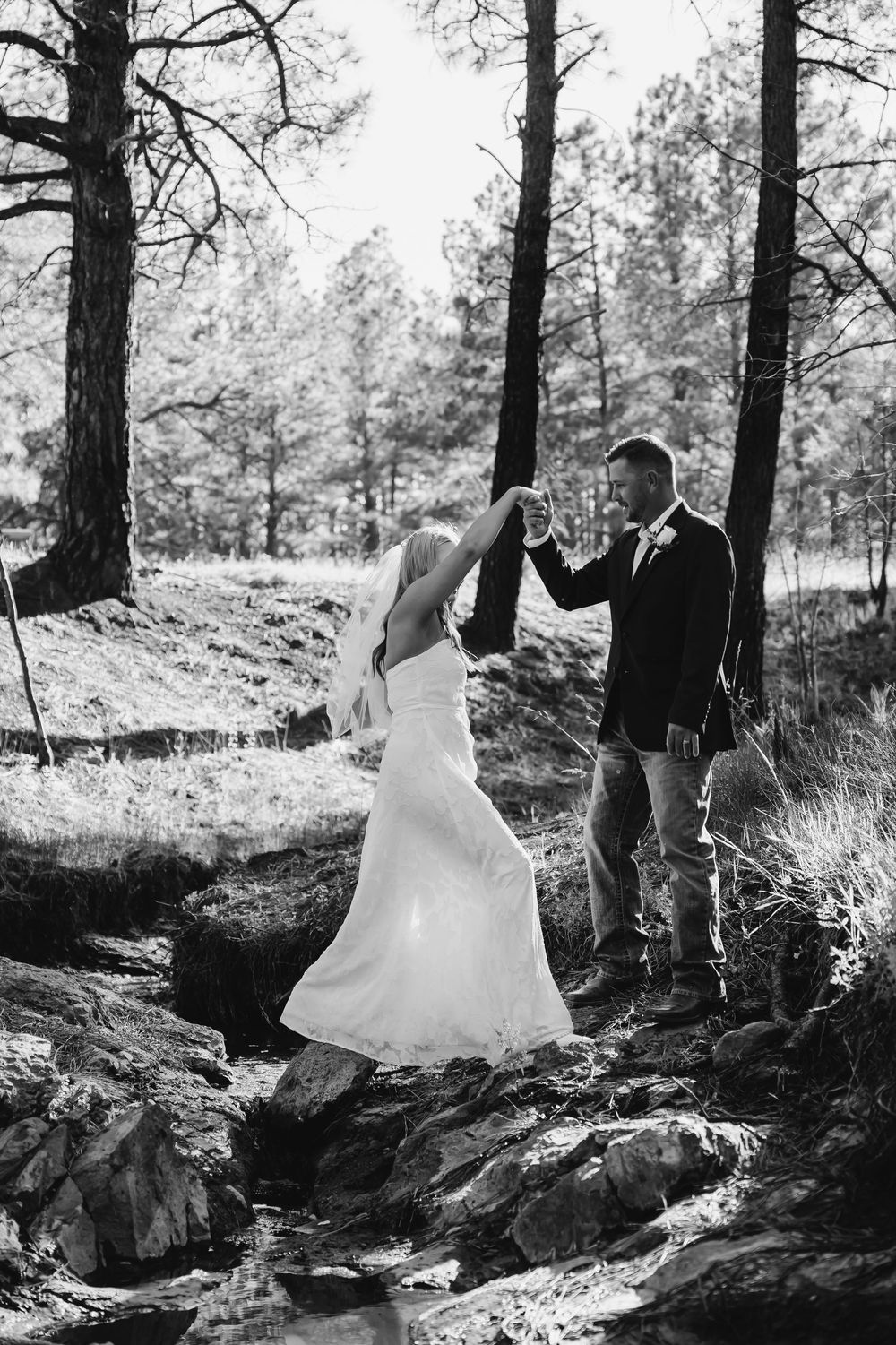 Black and white photo of bride and groom dancing among pine trees on rocky terrain during outdoor wedding.