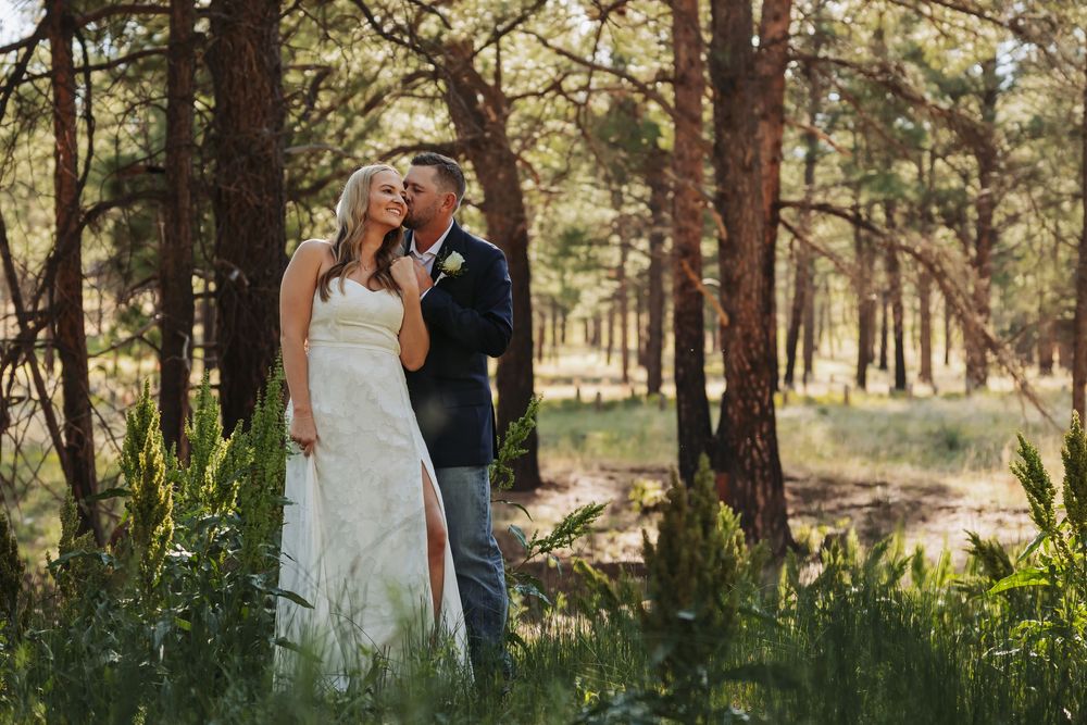 Bride and groom share a romantic moment in a sunlit pine forest, bride in white gown, groom in navy suit.