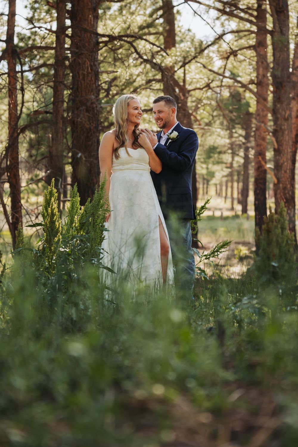 Bride and groom share a romantic moment among tall pine trees and lush greenery during an outdoor forest wedding.