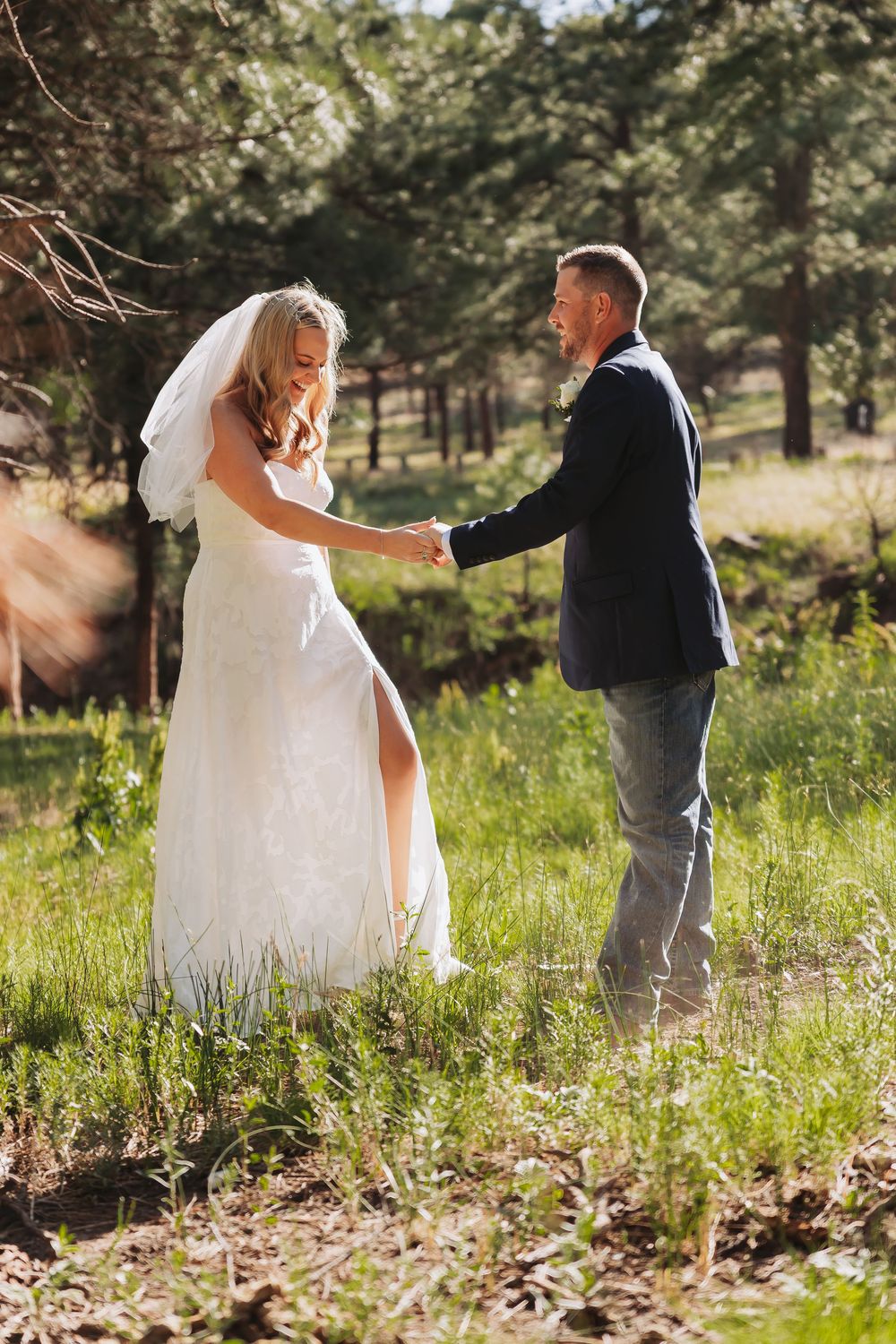 Bride in white gown and veil holds hands with groom in navy blazer and jeans in a sunlit forest meadow.