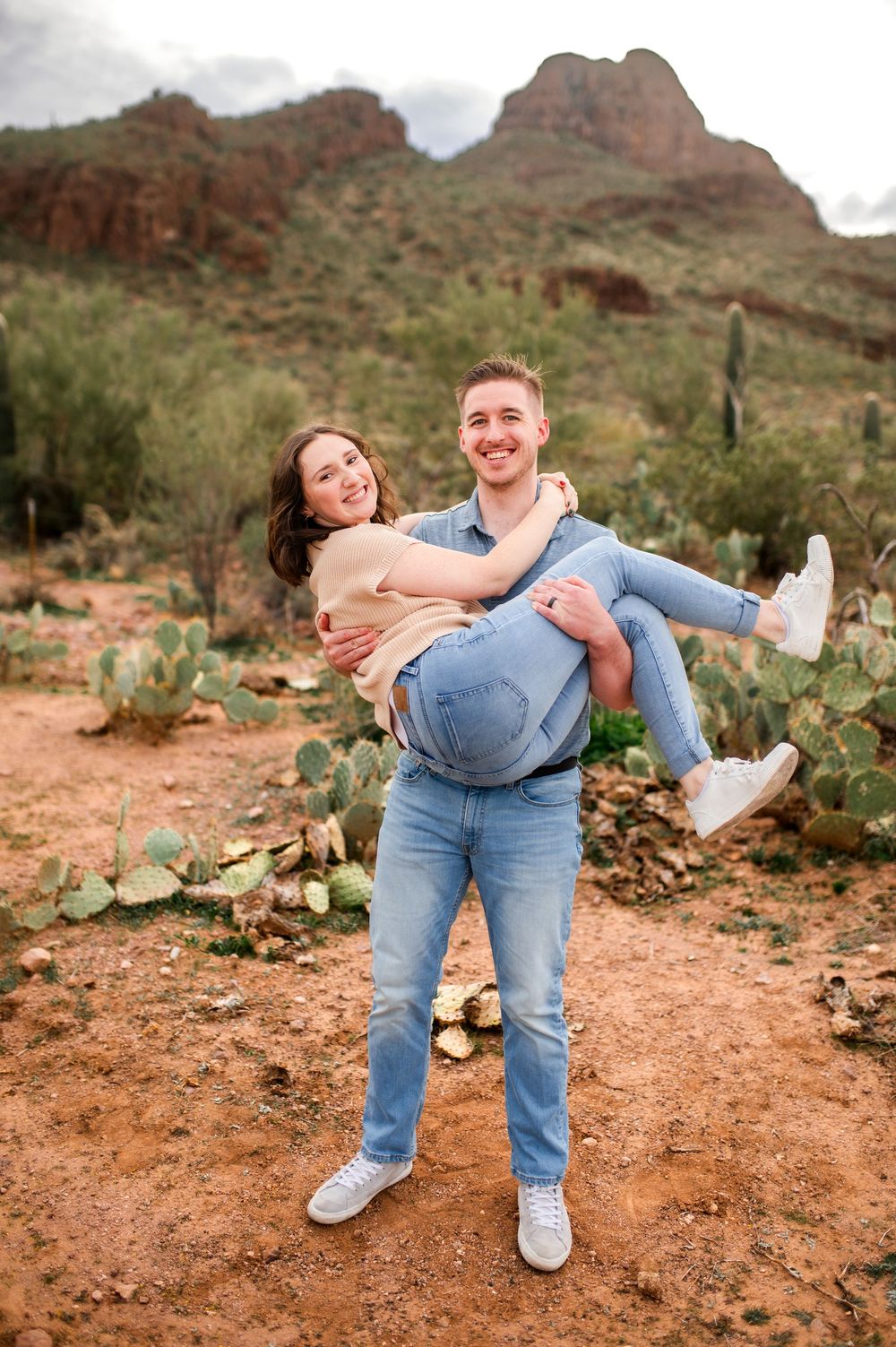 A joyful couple poses in the Arizona desert, man carrying woman in his arms with saguaro cacti and red mountains behind.
