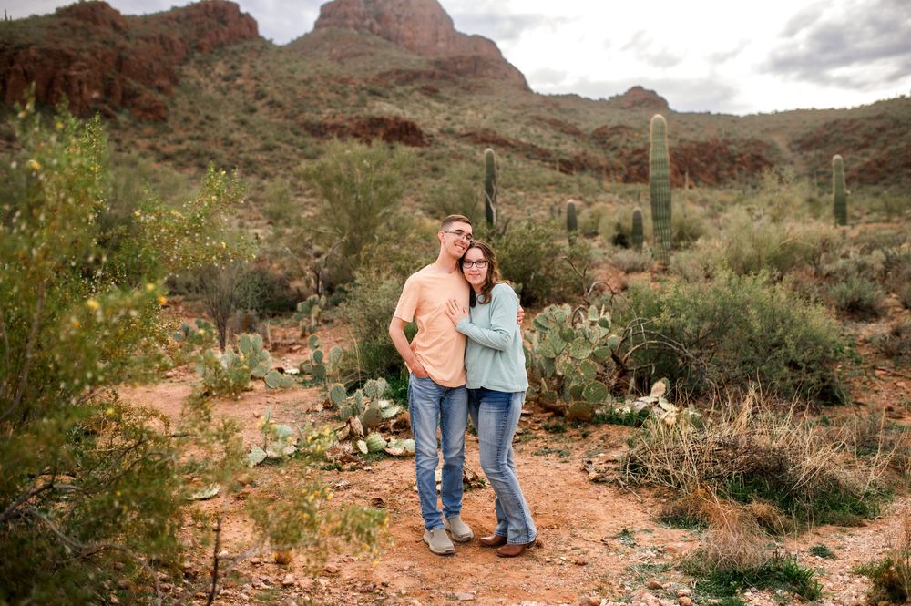 Smiling couple embraces on a desert trail surrounded by saguaro cacti and red rock mountains in Arizona.