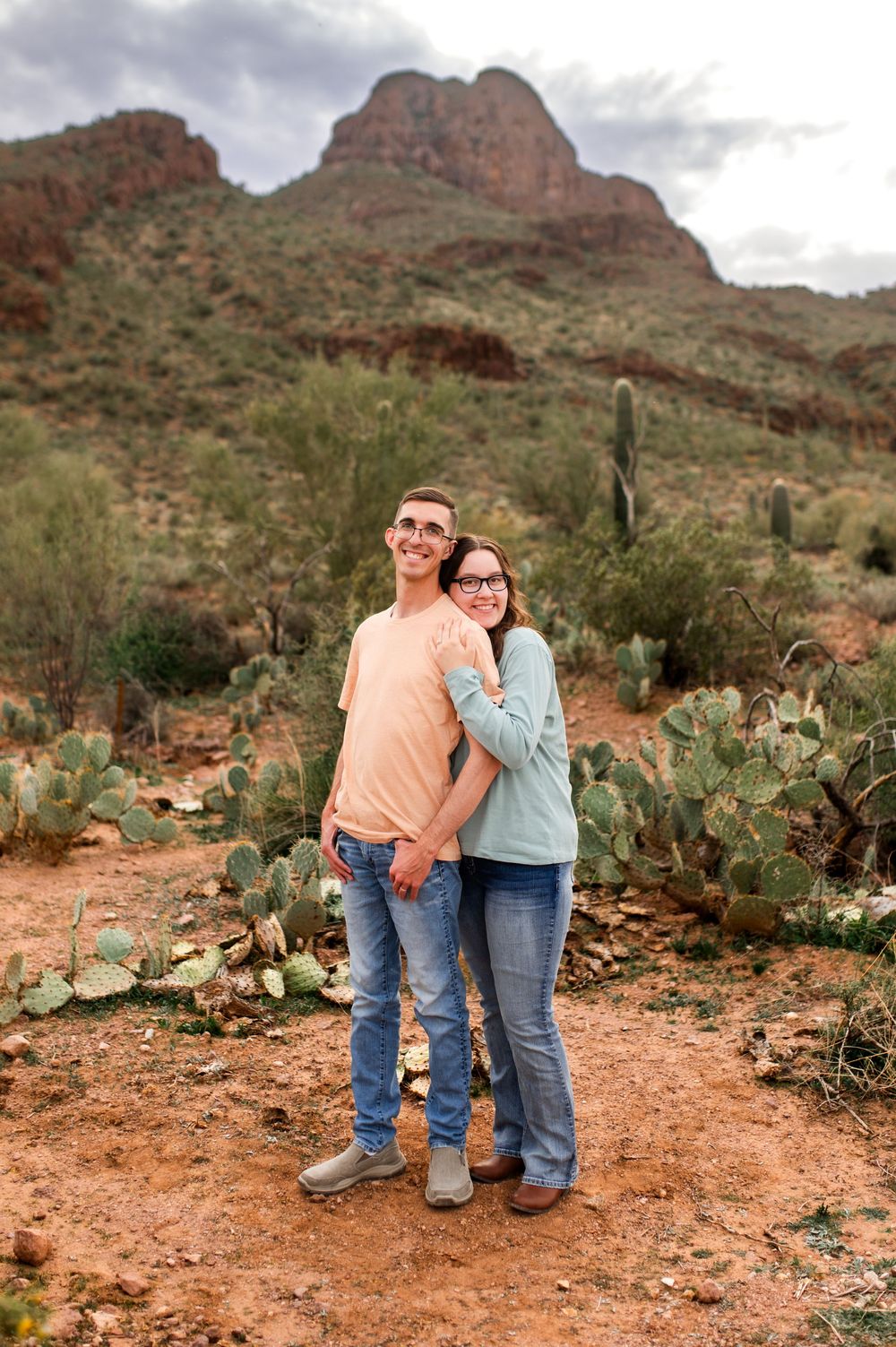 Happy couple holds hands in Arizona desert landscape with prickly pear cacti and dramatic red rock mountain backdrop.
