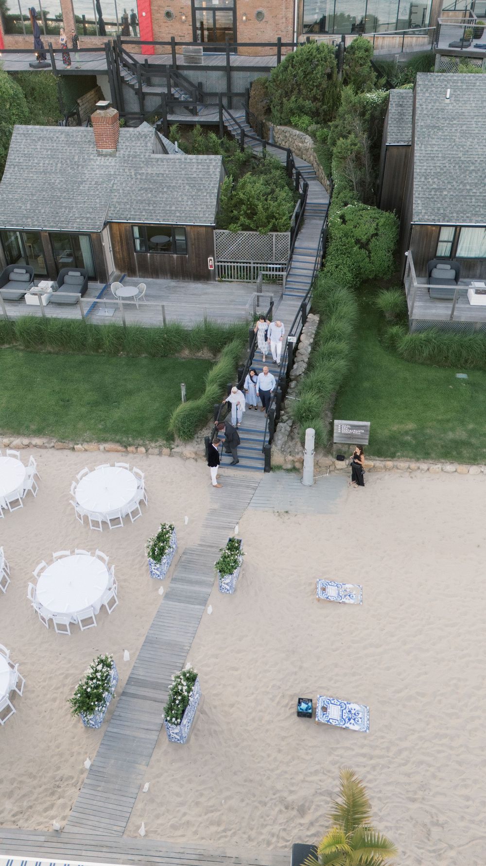 Aerial view of a beach wedding reception setup with white tables, floral arrangements, and guests walking on a wooden boardwalk.