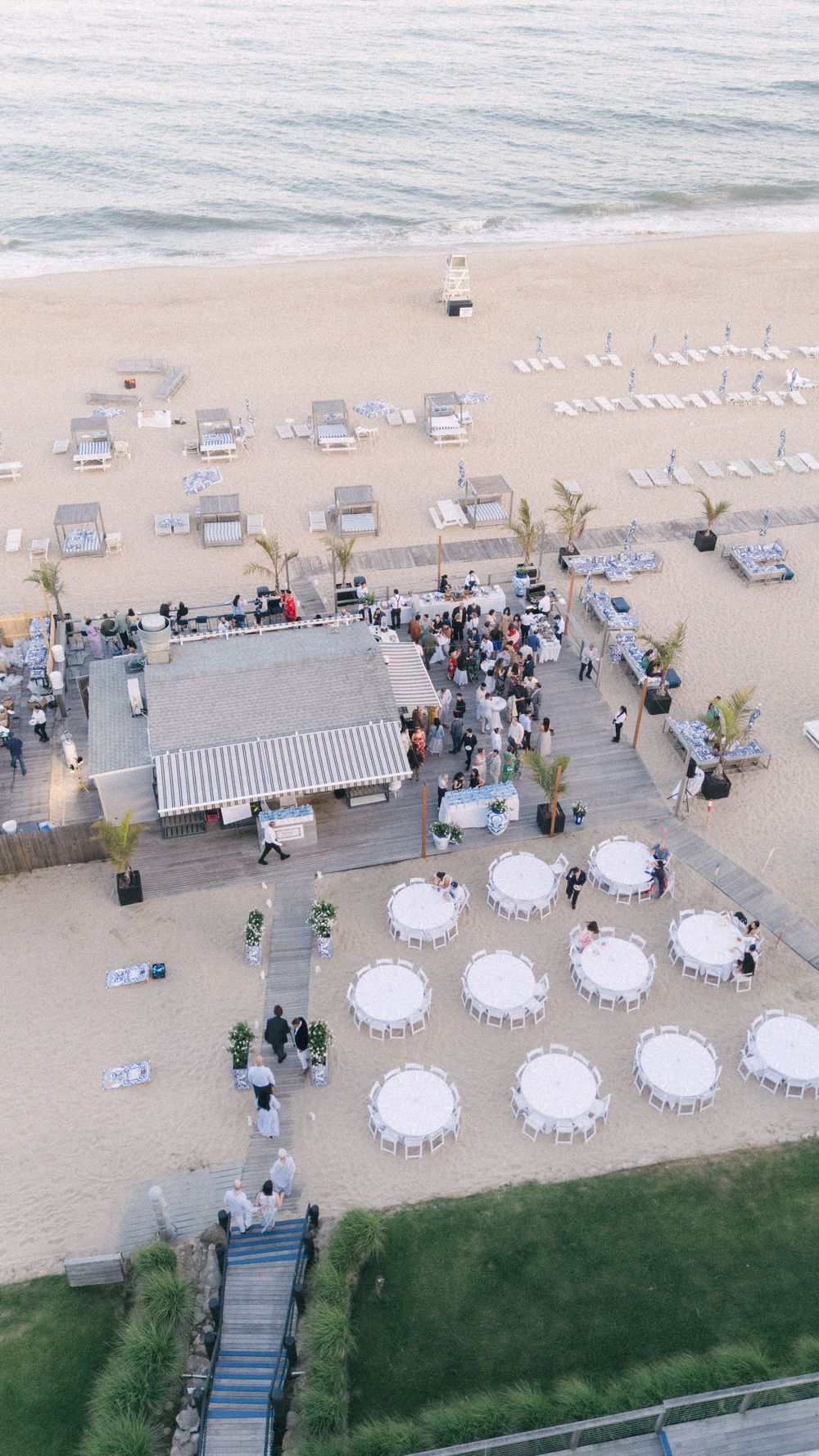 Aerial view of a beach wedding reception with round white tables, guests mingling on a wooden deck beside the ocean.