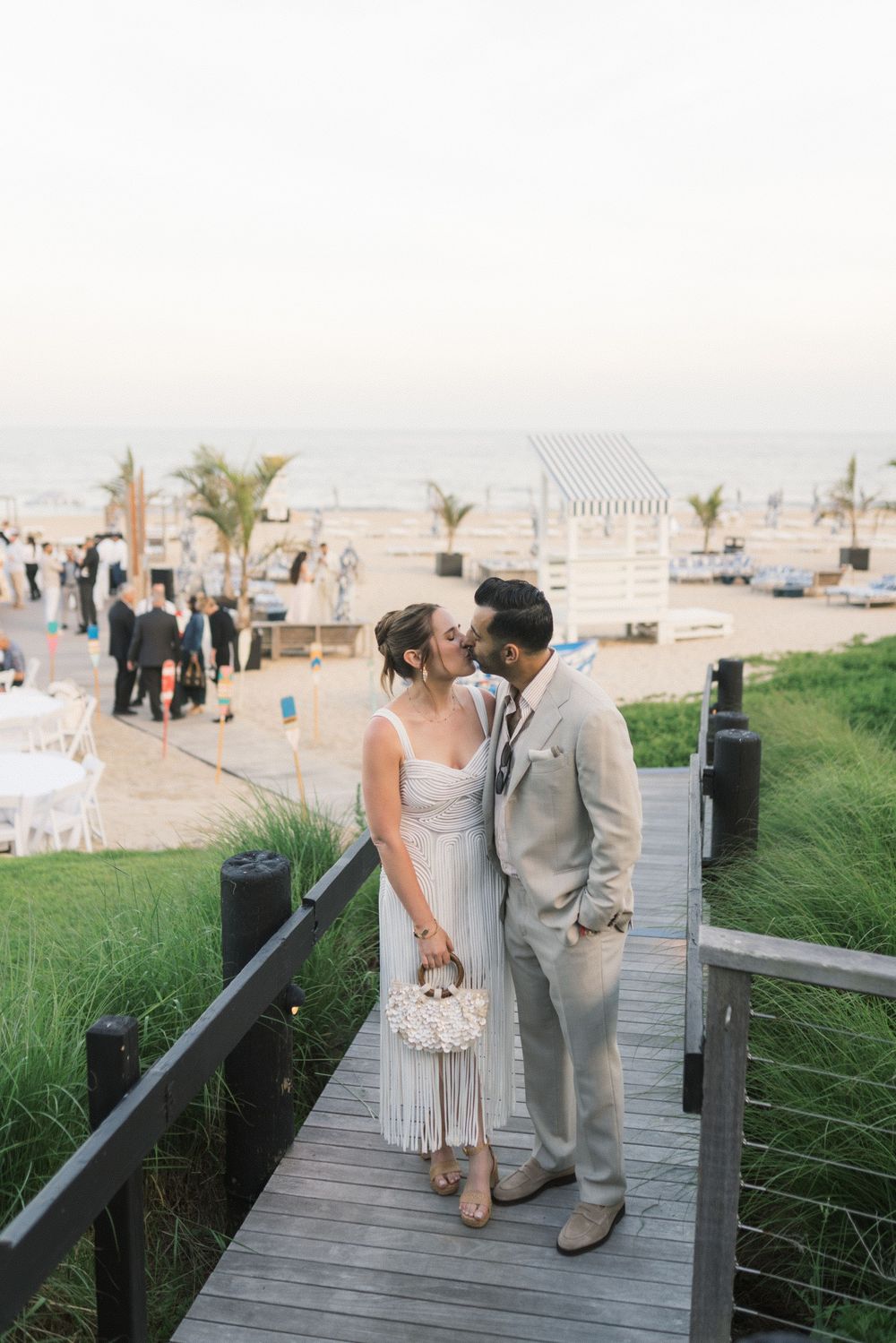 Couple sharing a kiss on a wooden boardwalk overlooking a beach wedding reception at sunset.