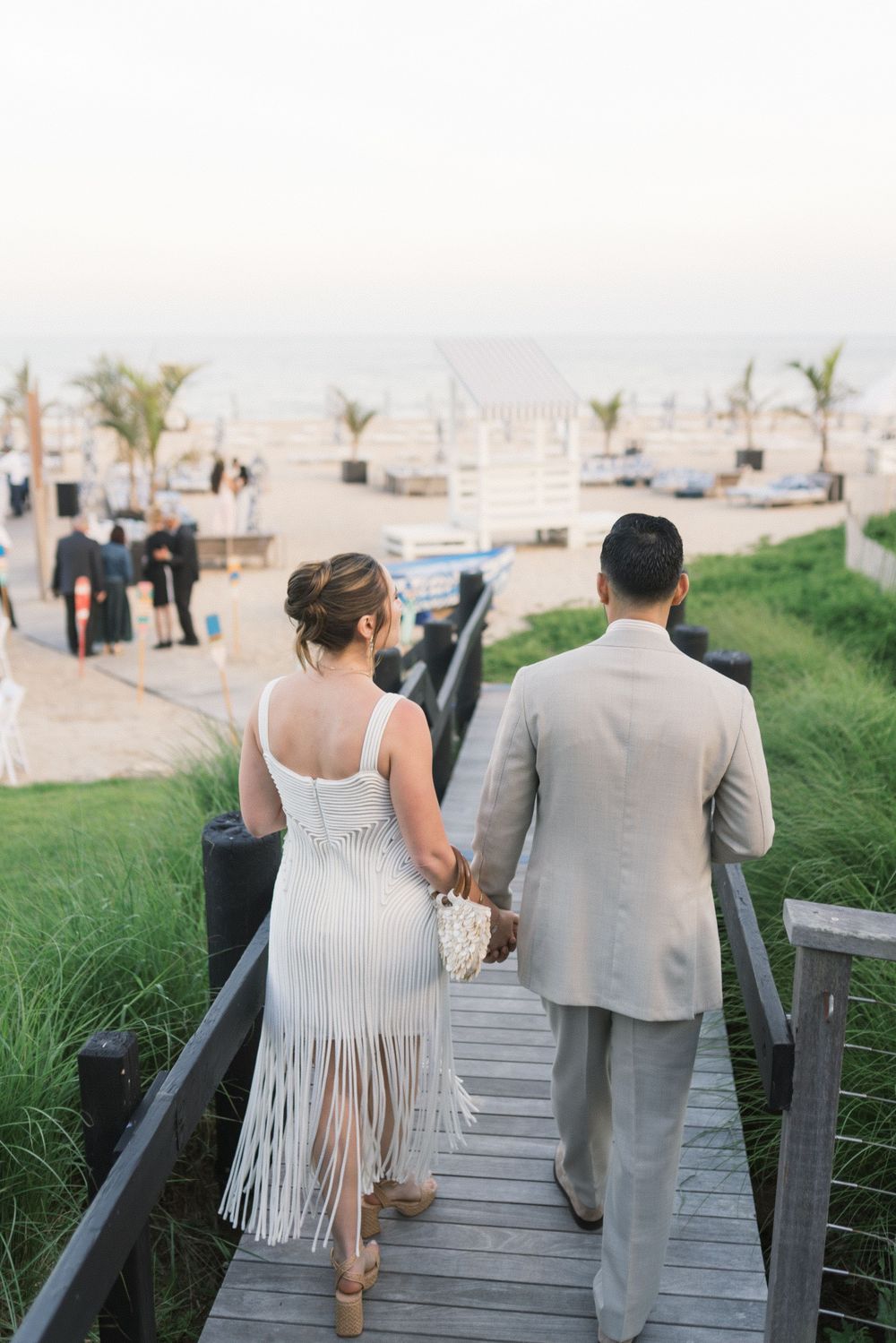 Couple in wedding attire walking hand-in-hand down a wooden boardwalk toward a beachside ceremony at sunset.