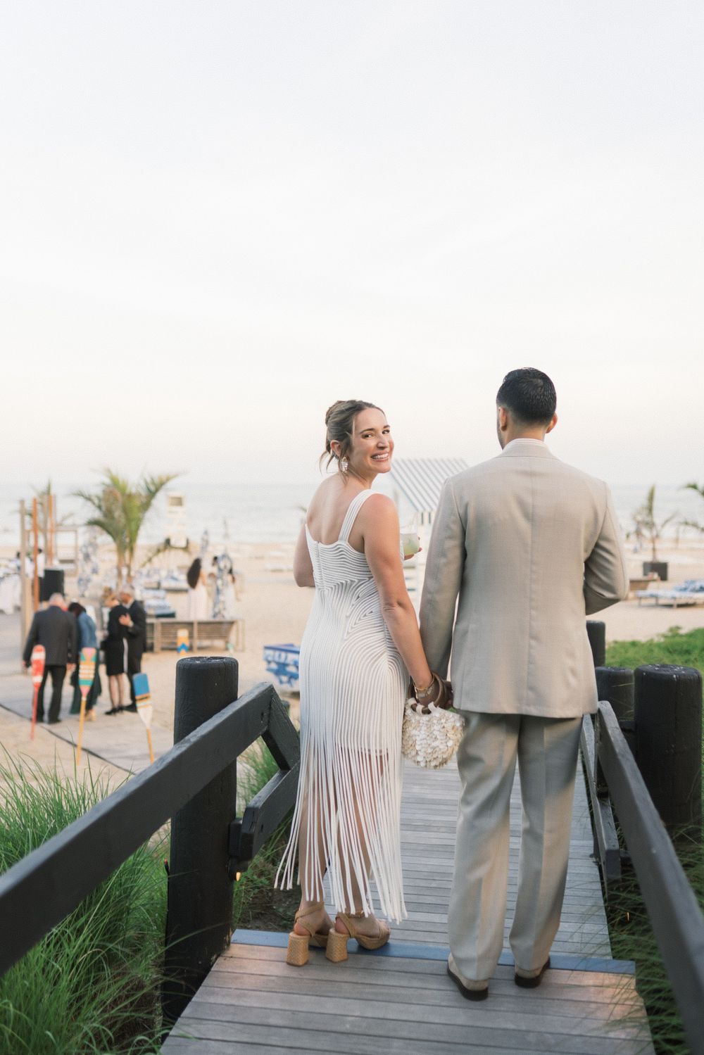 Bride in white fringe dress and groom in tan suit walking hand-in-hand on a beach boardwalk.