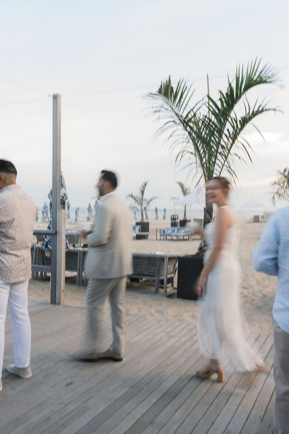 Elegantly dressed guests mingle on a beachside wooden deck at a twilight event.
