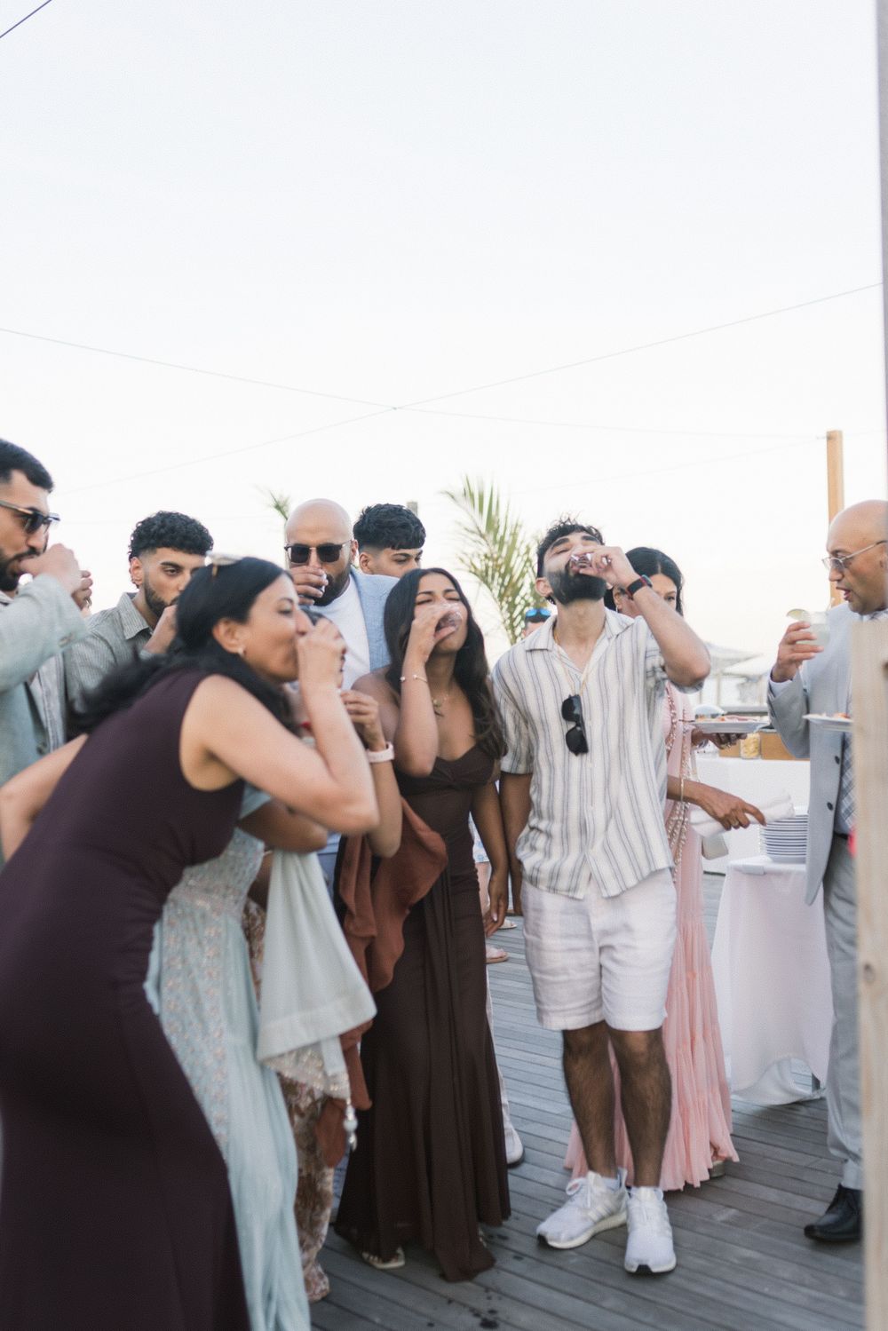 Guests laughing and drinking at an outdoor beach wedding cocktail hour on a wooden deck.
