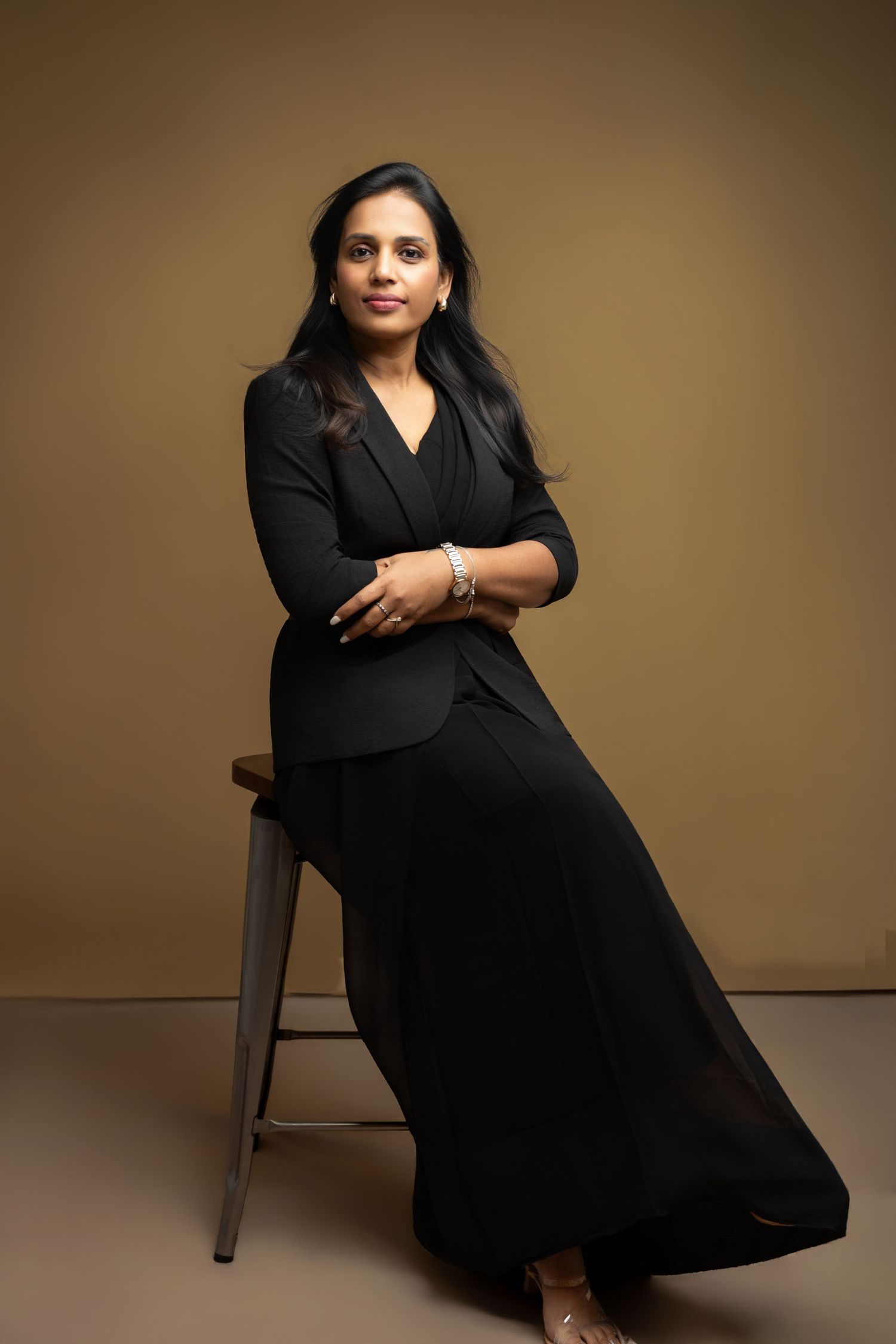 Woman in black blazer and skirt sits confidently on stool against warm brown background.