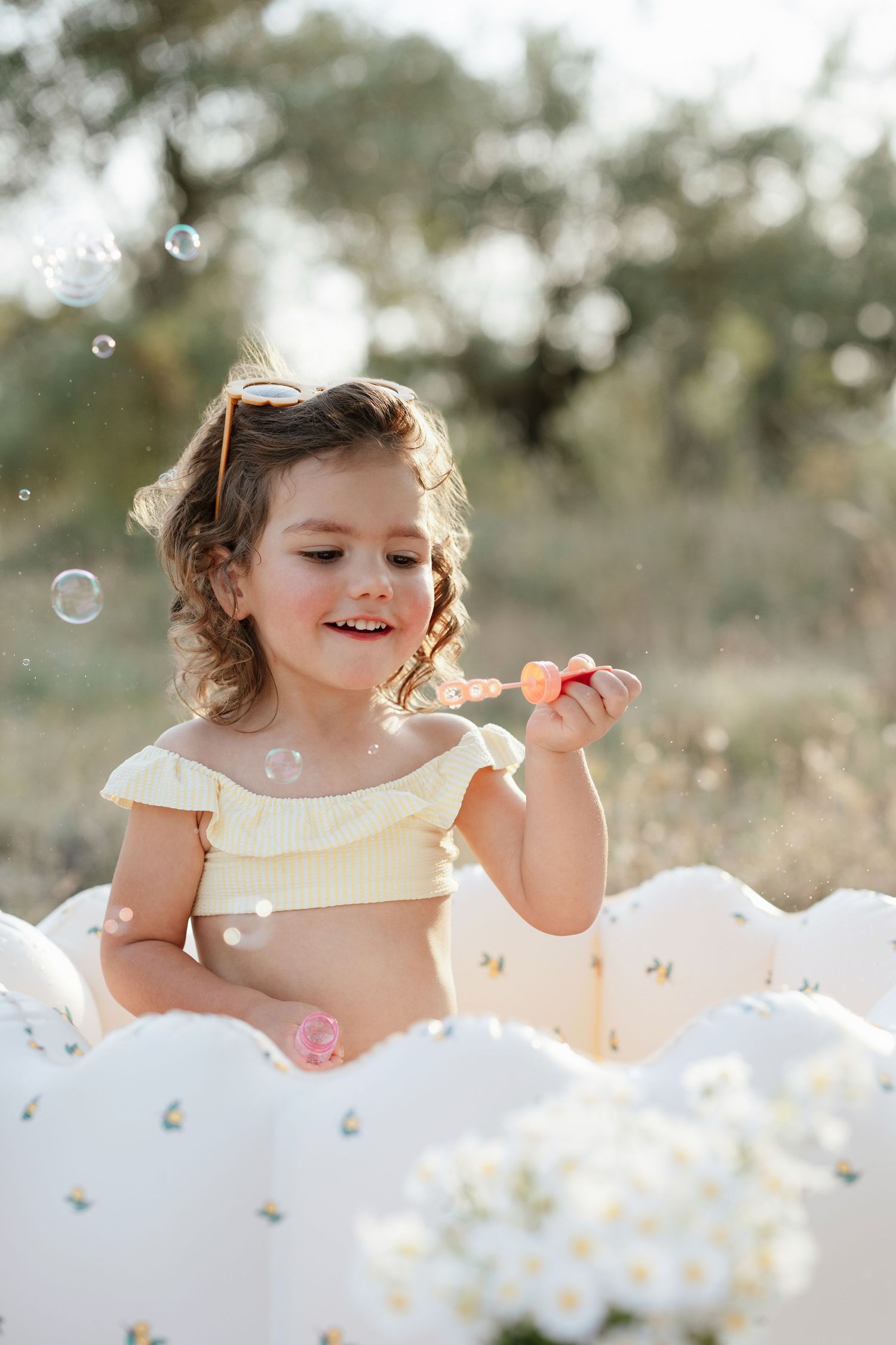 Young girl in yellow top blowing soap bubbles outdoors, surrounded by white balloons and flowers on sunny day.