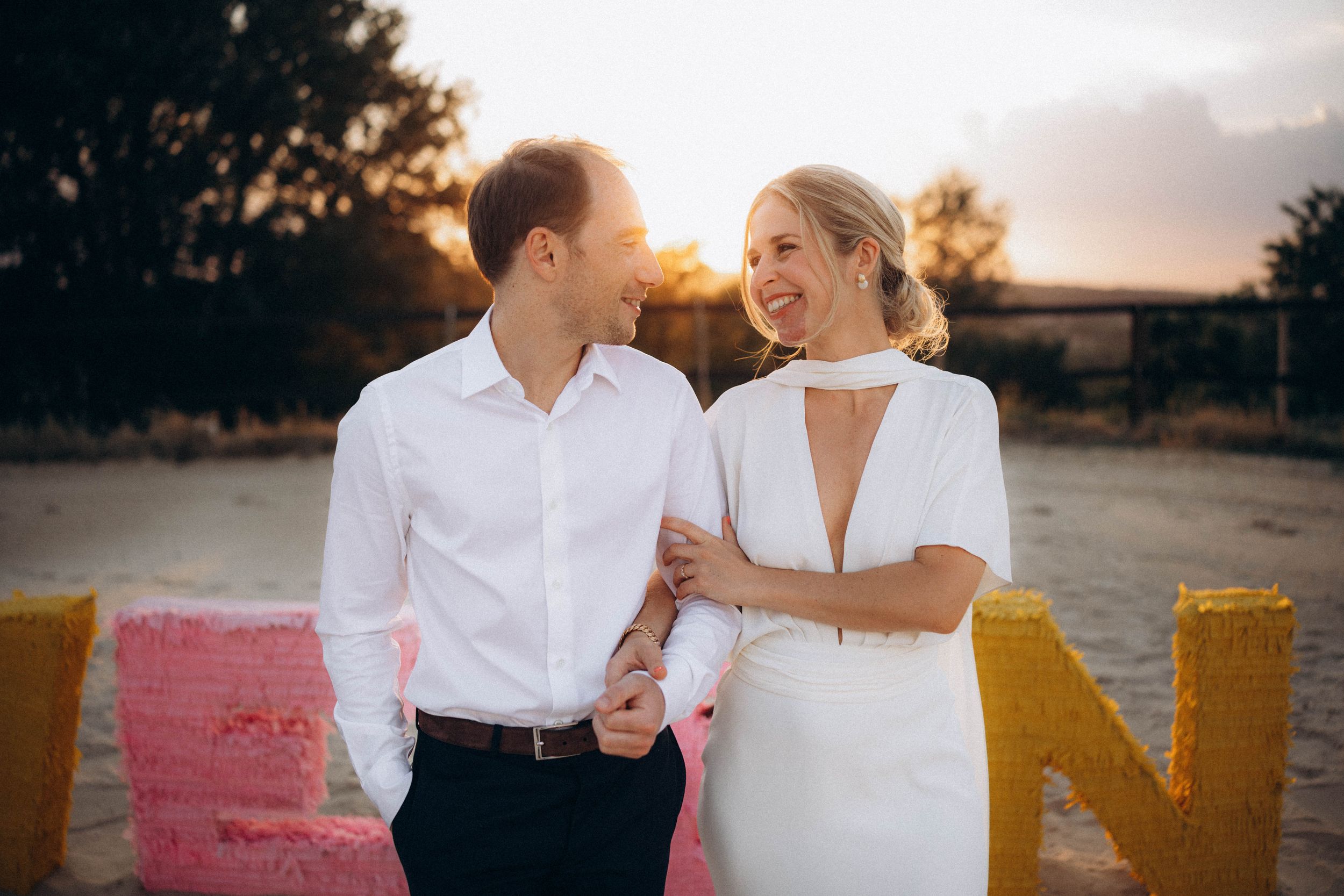 Couple in white attire holding hands at sunset with colorful letter decorations in background.
