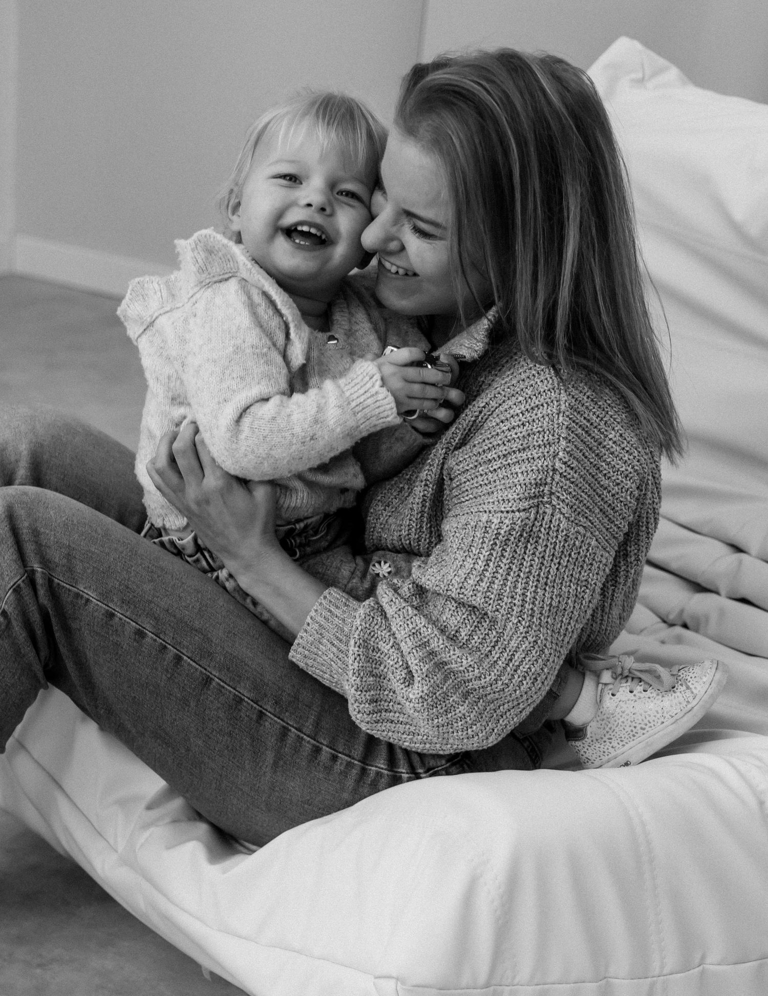 Black and white photo of a smiling mother holding her laughing baby girl while sitting on a bed.