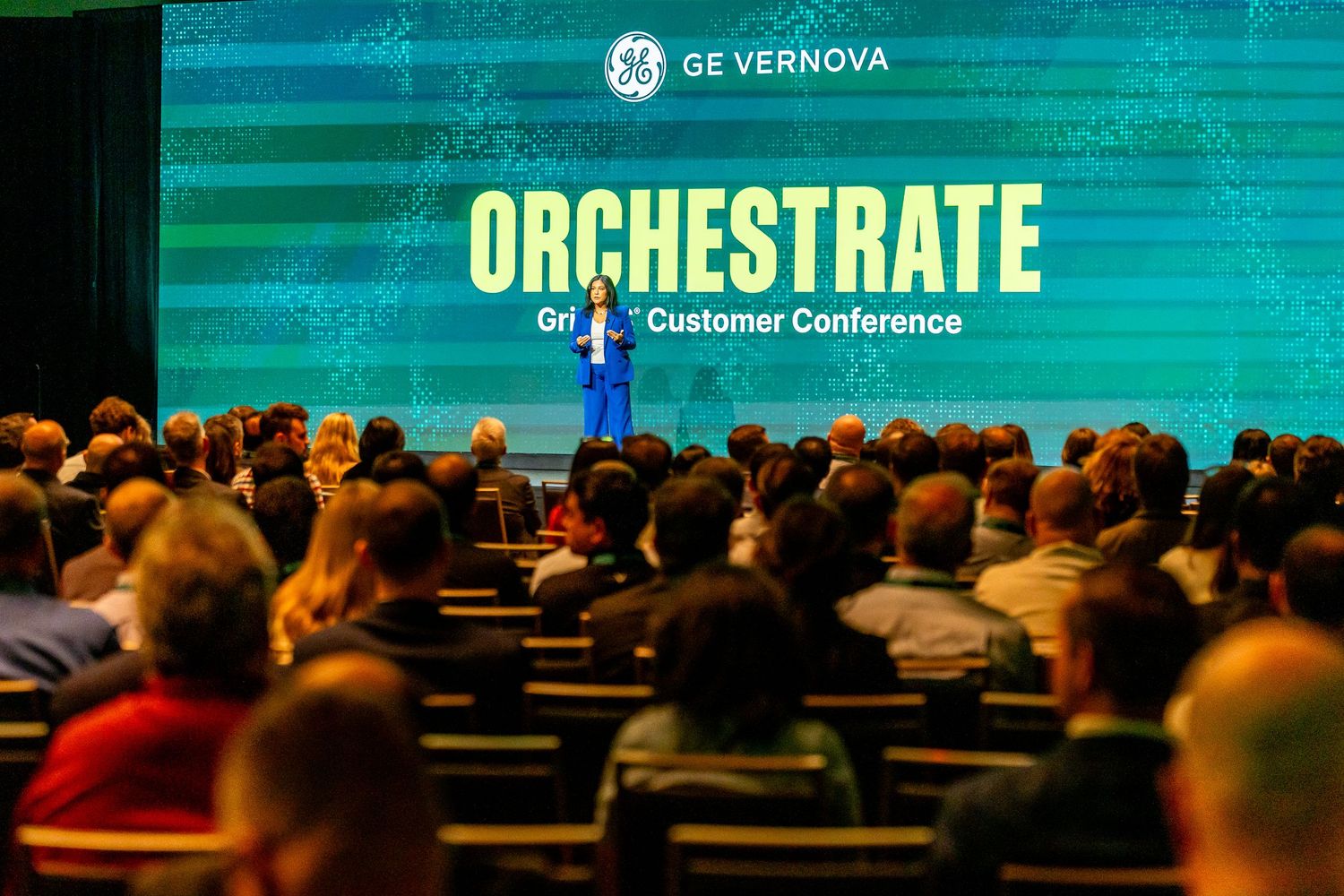 GE Vernova Orchestrate Grid Customer Conference presenter on stage addressing large audience in darkened conference hall.